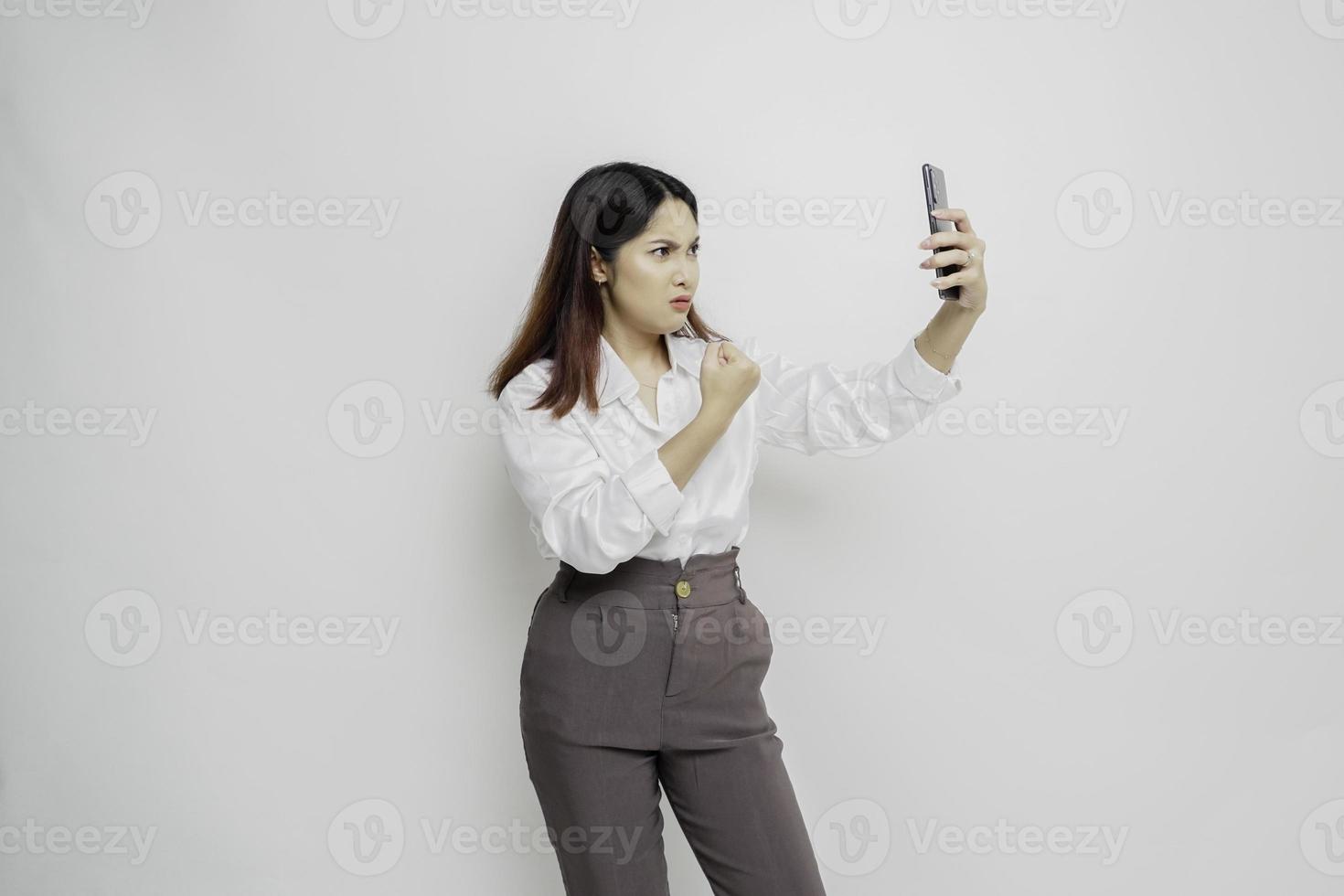 un' insoddisfatto giovane asiatico donna sembra scontento indossare bianca camicia irritata viso espressioni Tenere sua Telefono foto