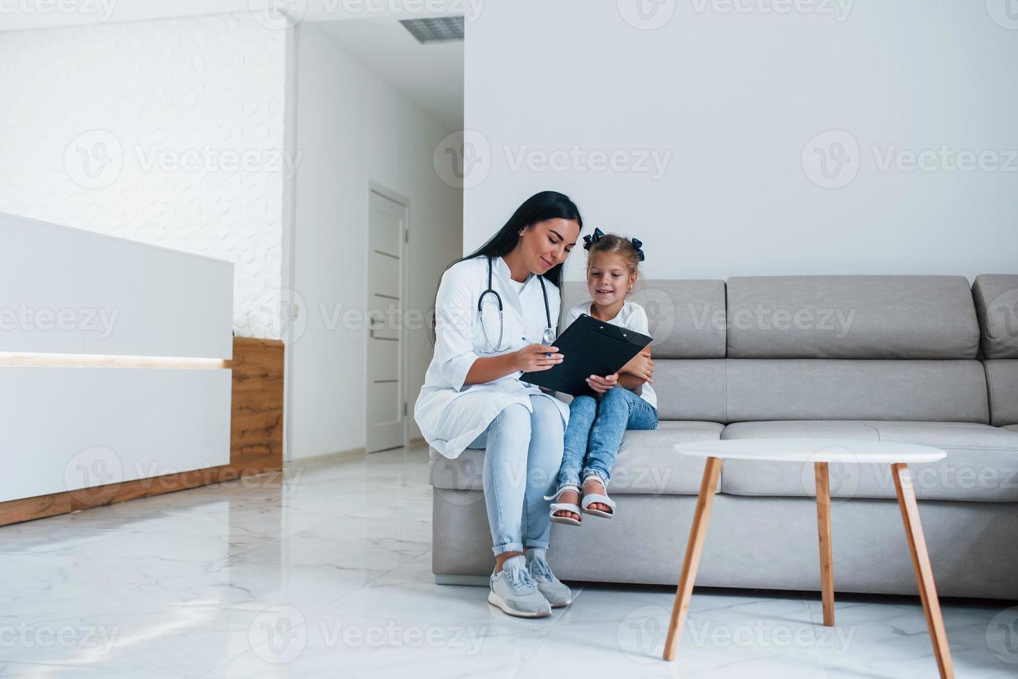 femmina medico con bloc notes si siede nel il clinica con poco ragazza foto