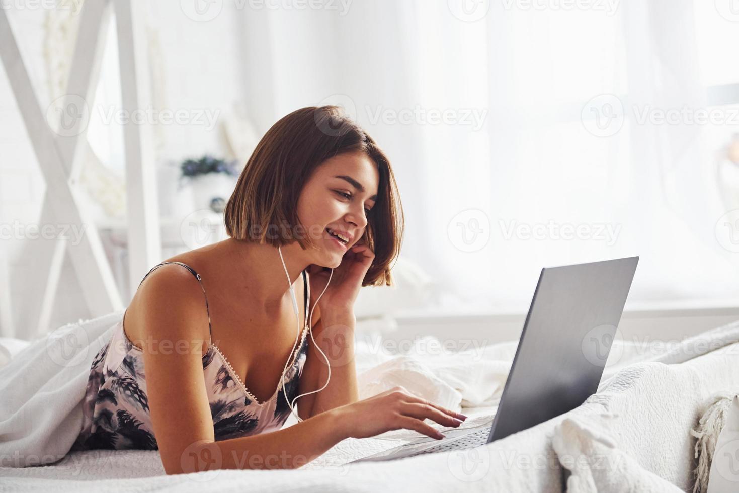 ascoltando per il musica di utilizzando cuffia. carino ragazza dire bugie su il letto con il computer portatile nel il Camera da letto a sua fine settimana tempo foto