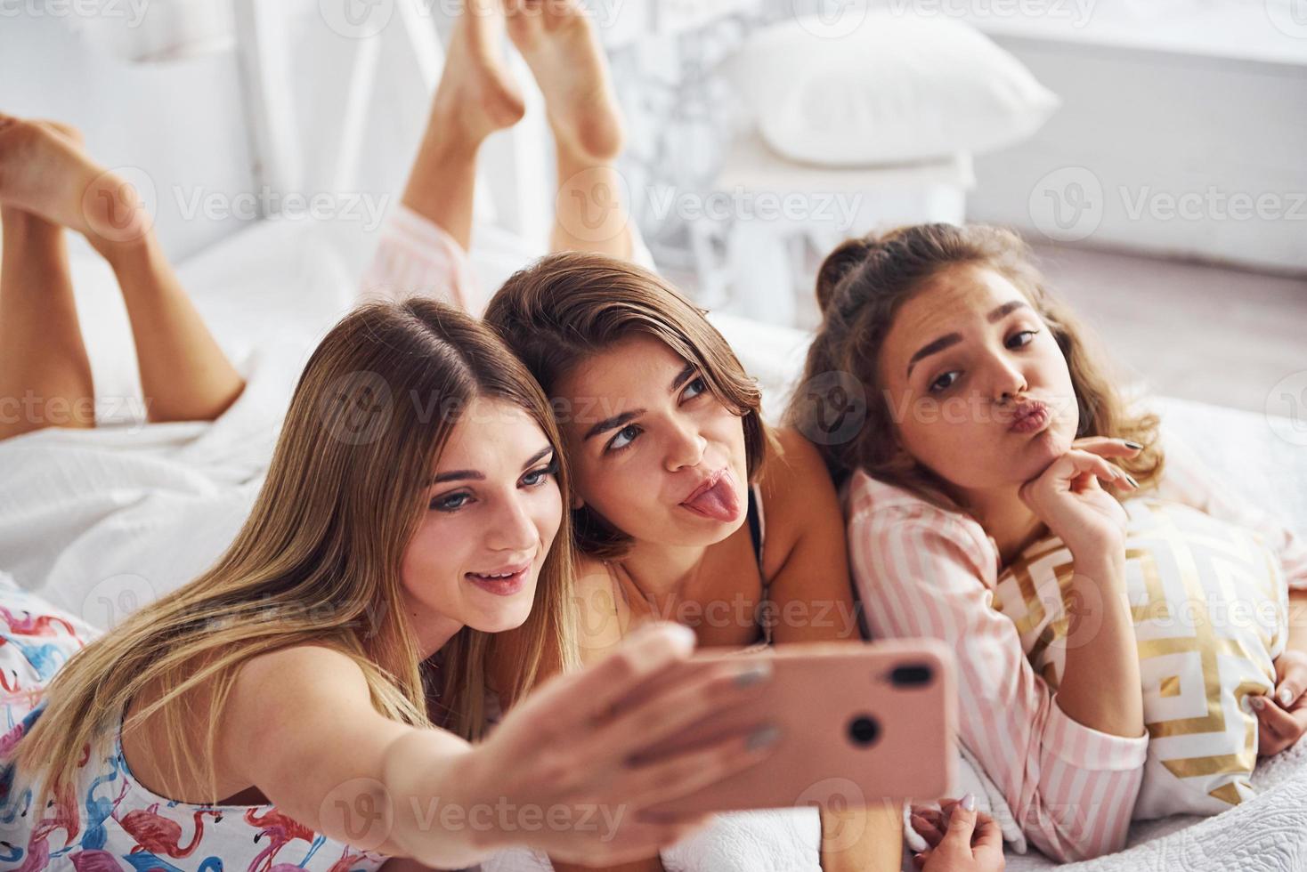 fabbricazione autoscatto di utilizzando rosa Telefono. contento femmina amici avendo bene tempo a pigiama festa nel il Camera da letto foto