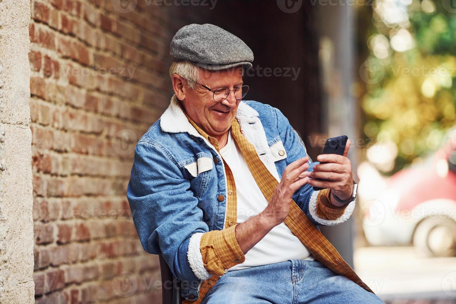 elegante anziano nel alla moda Abiti e nel bicchieri si siede su il sedia e usi Telefono foto