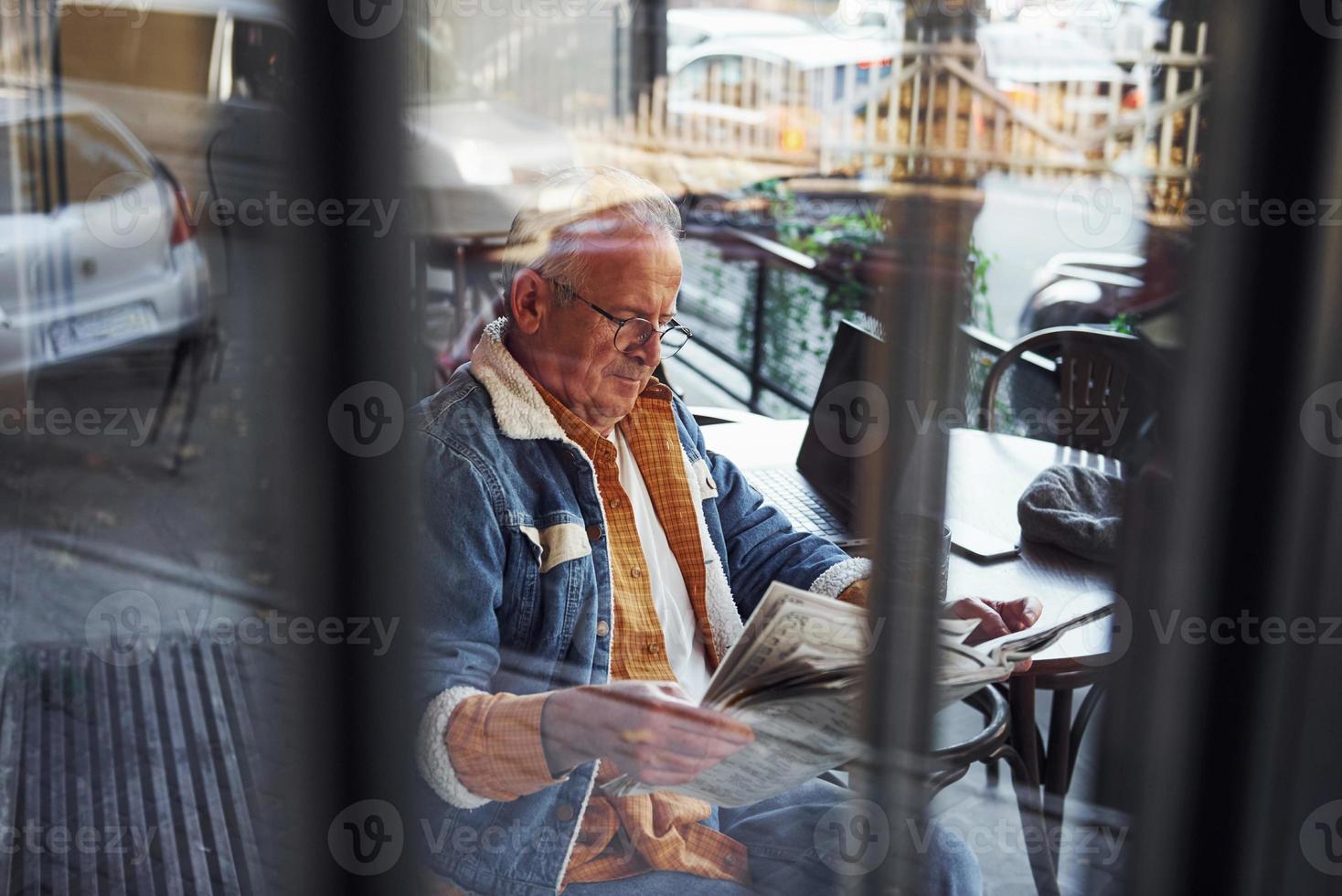 elegante anziano nel alla moda Abiti e nel bicchieri si siede nel il bar e legge giornale foto