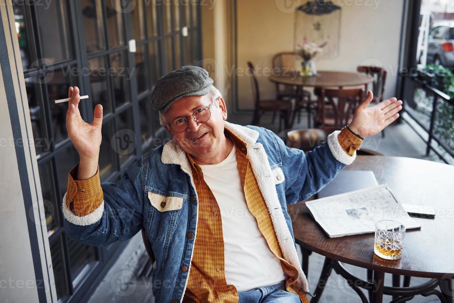 elegante anziano nel alla moda Abiti e nel bicchieri si siede nel il bar con sigaretta e parla foto