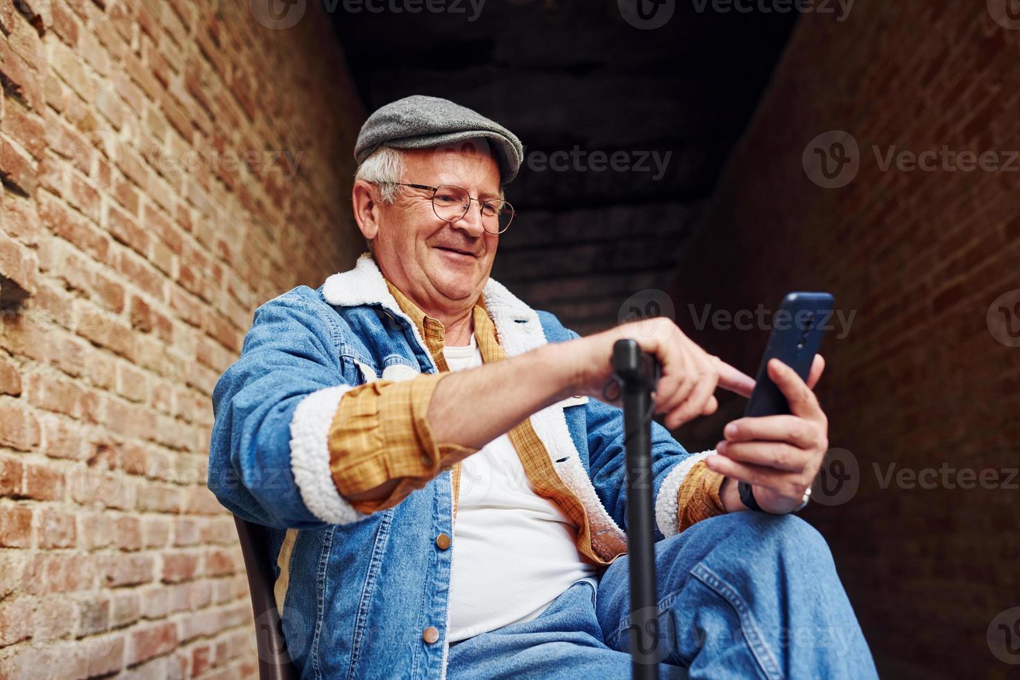 elegante anziano nel alla moda Abiti e nel bicchieri si siede su il sedia e usi Telefono foto