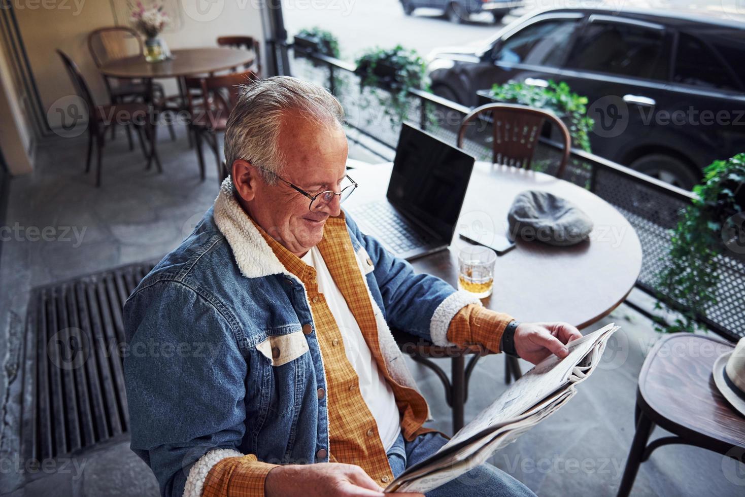 elegante anziano nel alla moda Abiti e nel bicchieri si siede nel il bar e legge giornale foto