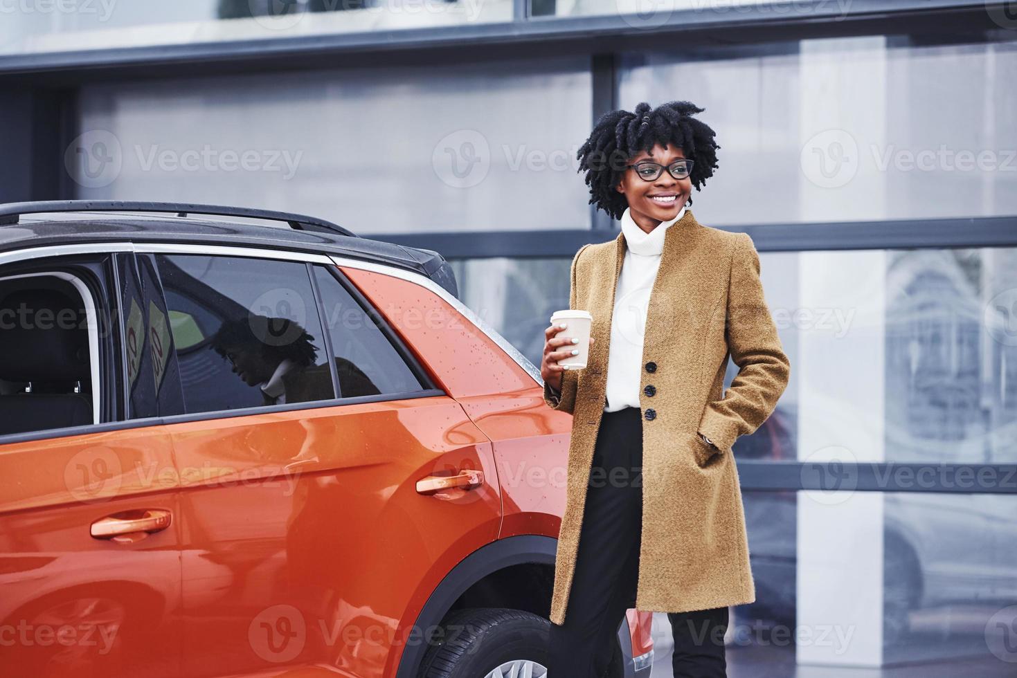 giovane africano americano donna nel bicchieri e con tazza di bevanda sta all'aperto vicino moderno auto foto