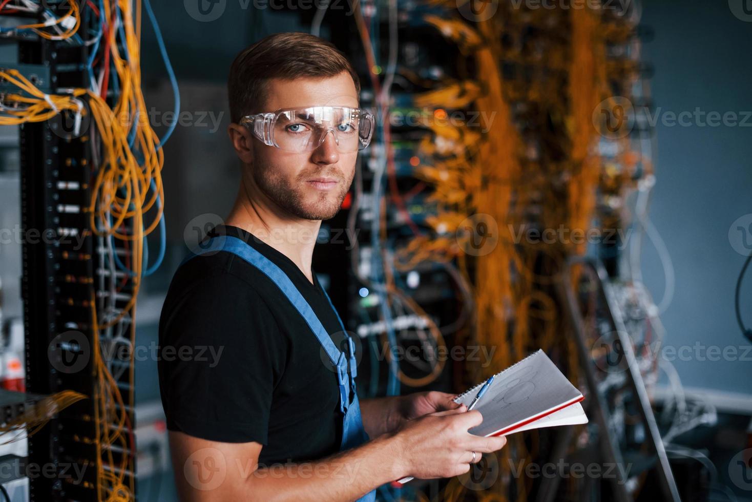 giovane uomo nel uniforme con bloc notes nel mani lavori con Internet attrezzatura e fili nel server camera foto