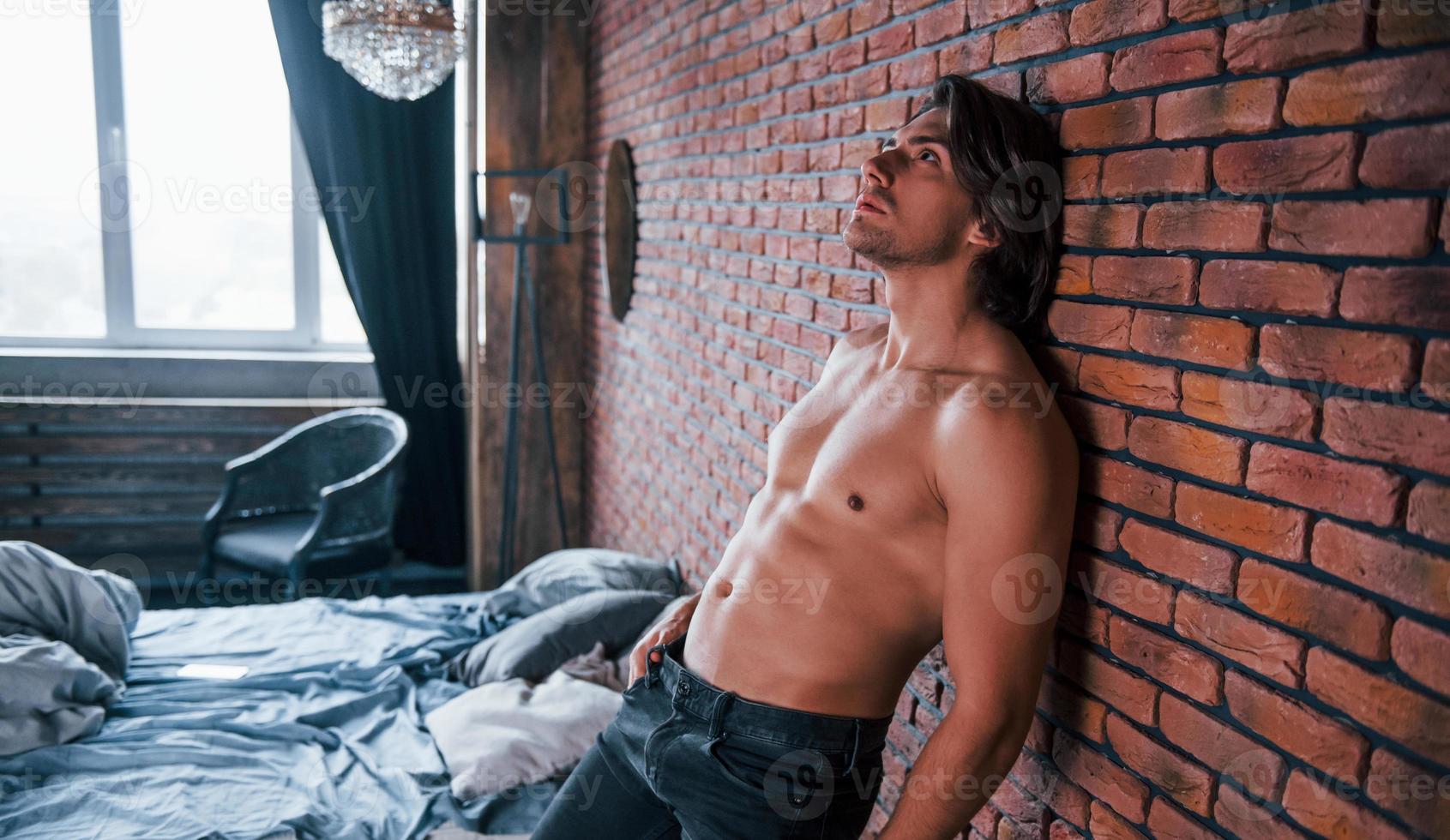 A petto nudo sexy uomo pendente su il mattone parete nel Camera da letto a mattina tempo foto