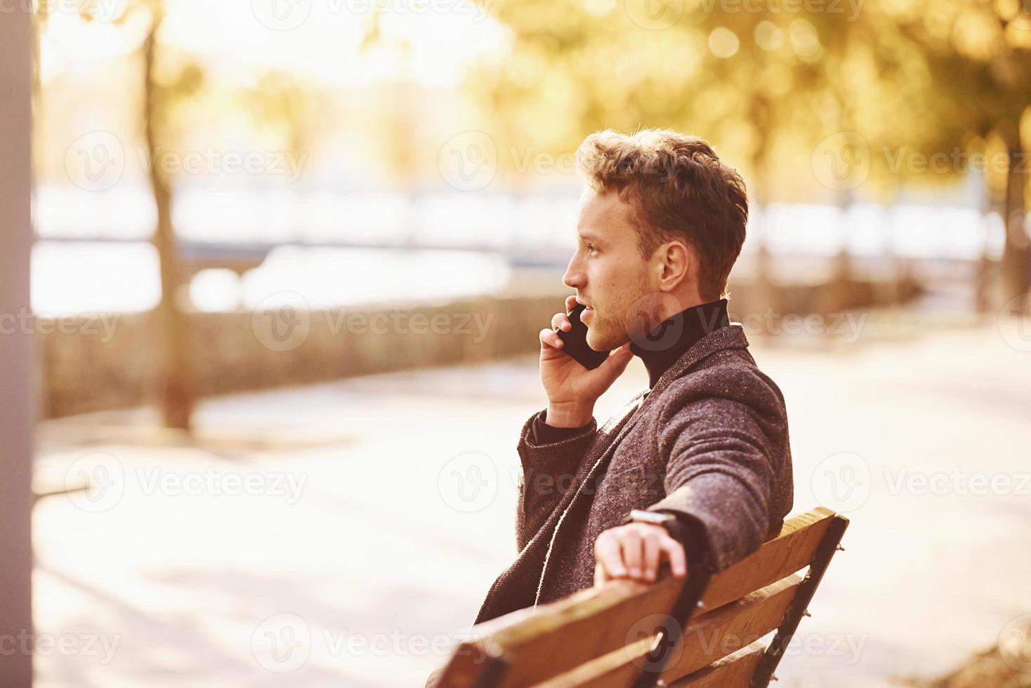 giovane uomo nel formale Abiti è nel il autunno parco a soleggiato giorno avere un' chiamata foto
