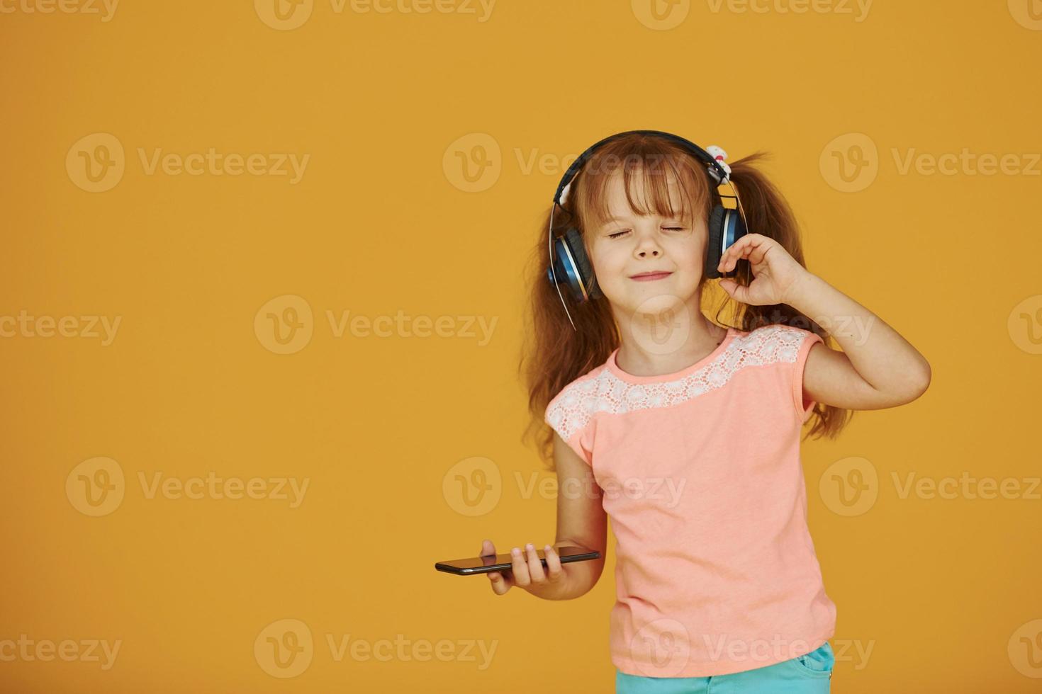 ritratto di carino poco ragazza nel cuffie e con Telefono nel il studio contro giallo sfondo foto