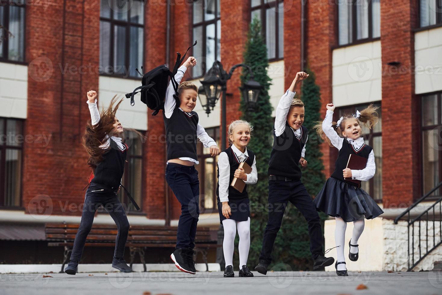 gruppo di bambini nel scuola uniforme salto e avendo divertimento all'aperto insieme vicino formazione scolastica edificio foto