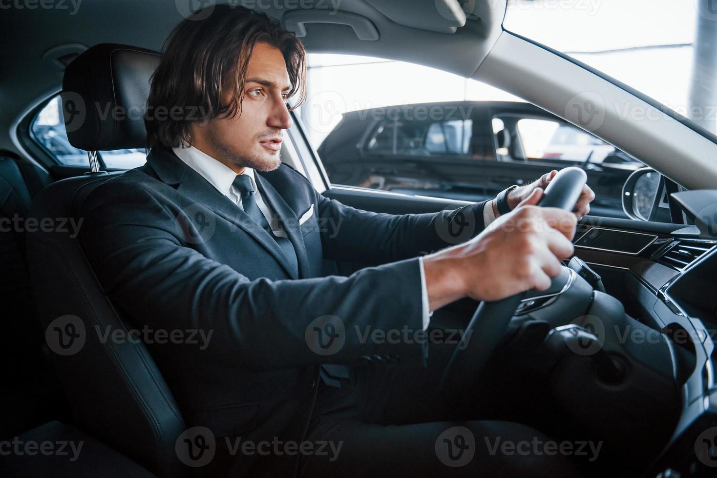 giovane uomo d'affari nel nero completo da uomo e cravatta guida moderno automobile foto