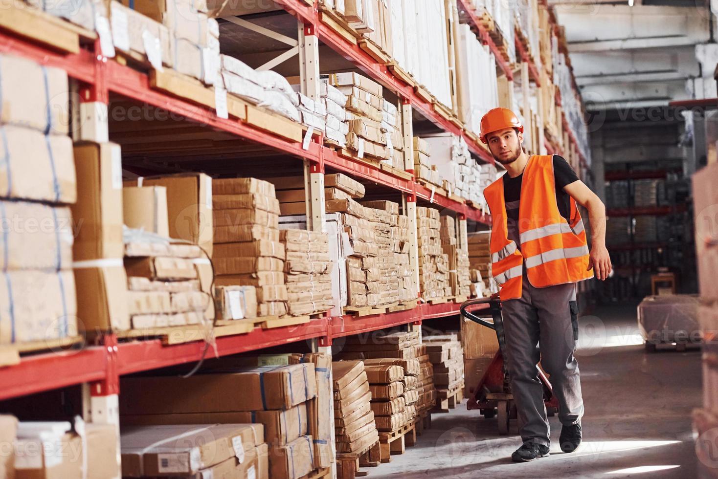 giovane maschio lavoratore nel uniforme è nel il magazzino spingendo pallet camion foto