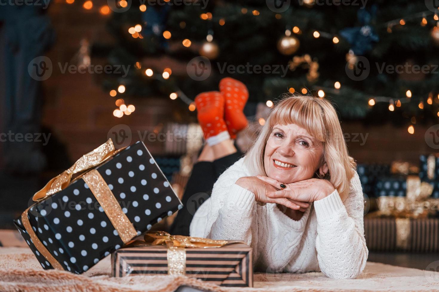 anziano allegro donna dire bugie giù nel il camera con Natale regalo scatole foto