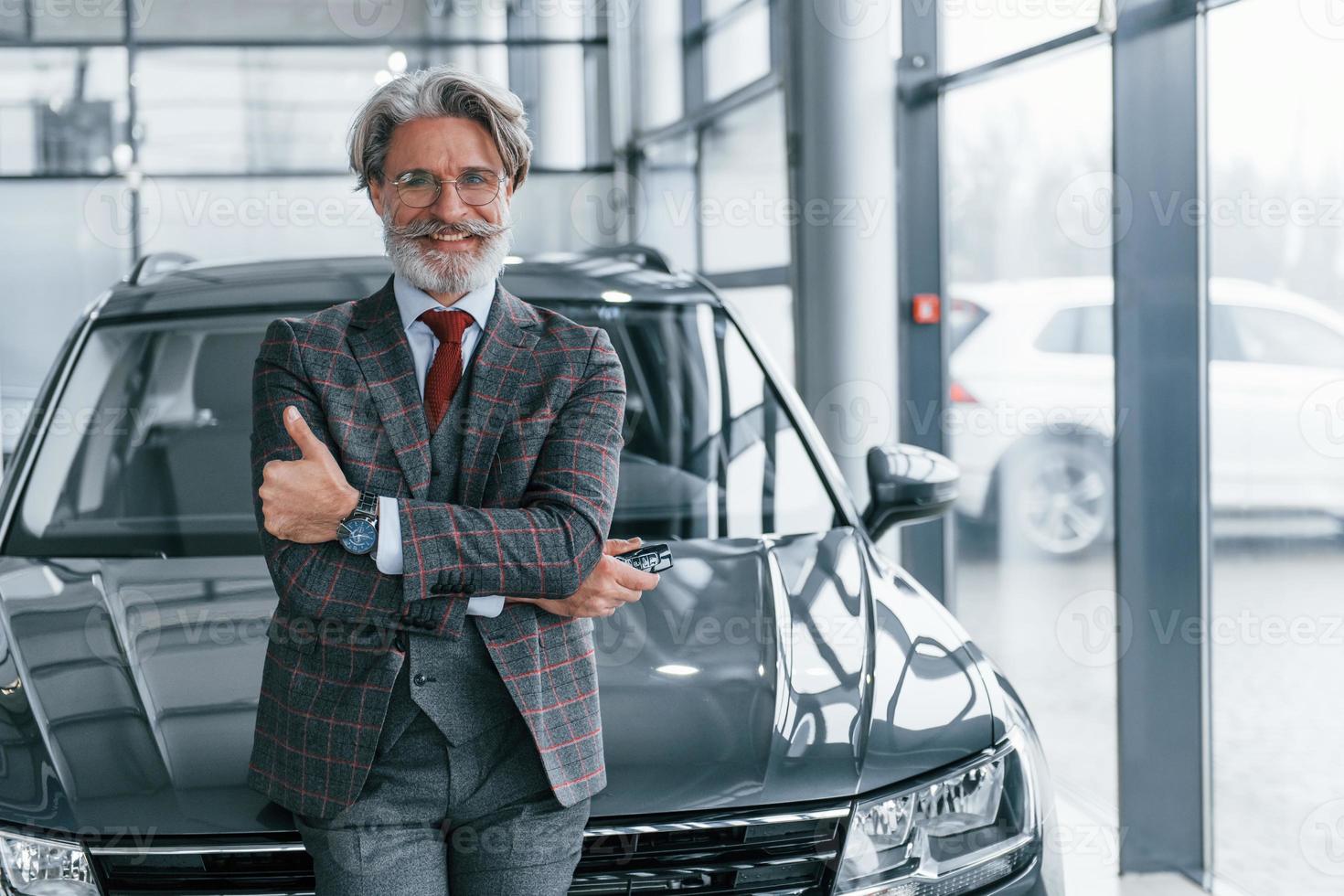 anziano uomo d'affari nel completo da uomo e cravatta con grigio capelli e barba in piedi in casa vicino auto foto