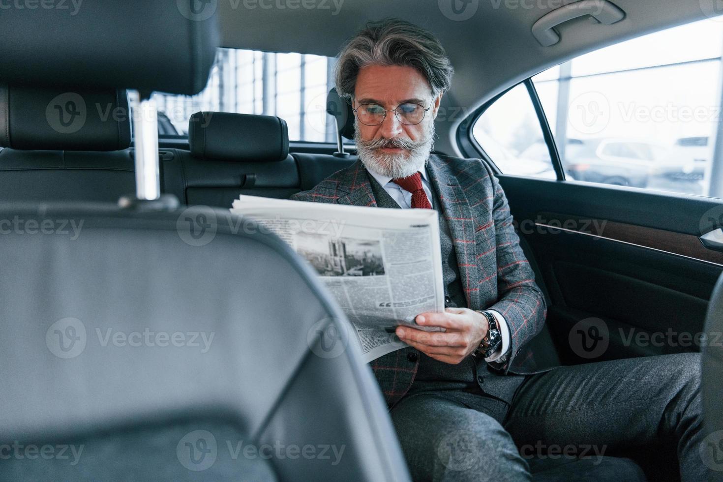 moderno elegante anziano uomo con grigio capelli e baffi lettura giornale dentro di il auto foto