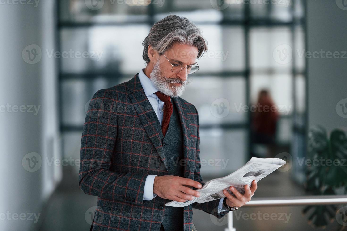 anziano uomo d'affari nel completo da uomo e cravatta con grigio capelli e barba detiene giornale nel mani foto