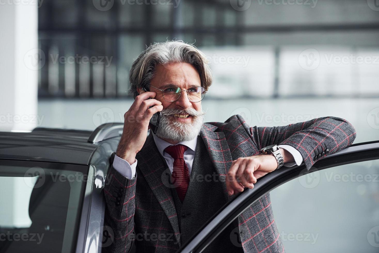 anziano uomo d'affari nel completo da uomo e cravatta con grigio capelli e barba è vicino auto parlando di il Telefono e sorridente foto