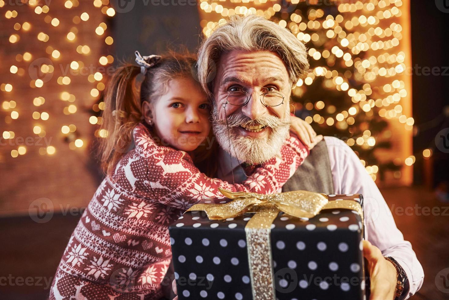 allegro stile anziano uomo con grigio capelli e barba seduta con poco ragazza nel decorato Natale camera foto