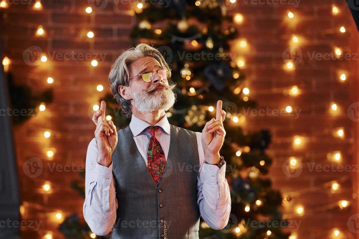 ritratto di elegante anziano con grigio capelli e barba nel giacca e bianca camicia è nel decorato Natale camera foto