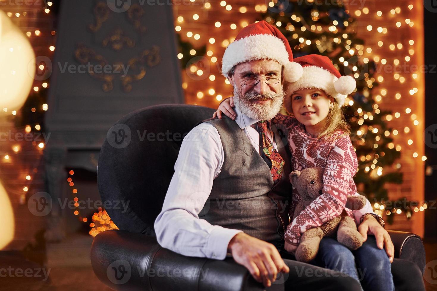 allegro stile anziano uomo con grigio capelli e barba seduta con poco ragazza nel Natale cappelli e orsacchiotto orso foto