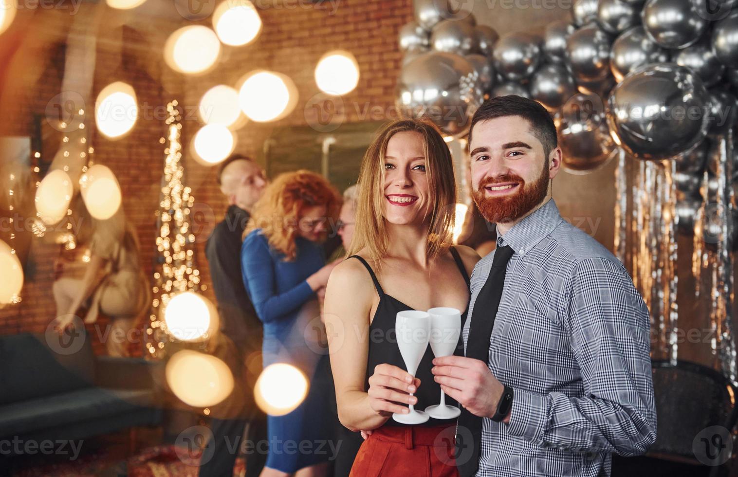 barbuto giovane uomo con il suo fidanzata in piedi insieme contro loro amici nel Natale decorato camera e festeggiare nuovo anno foto