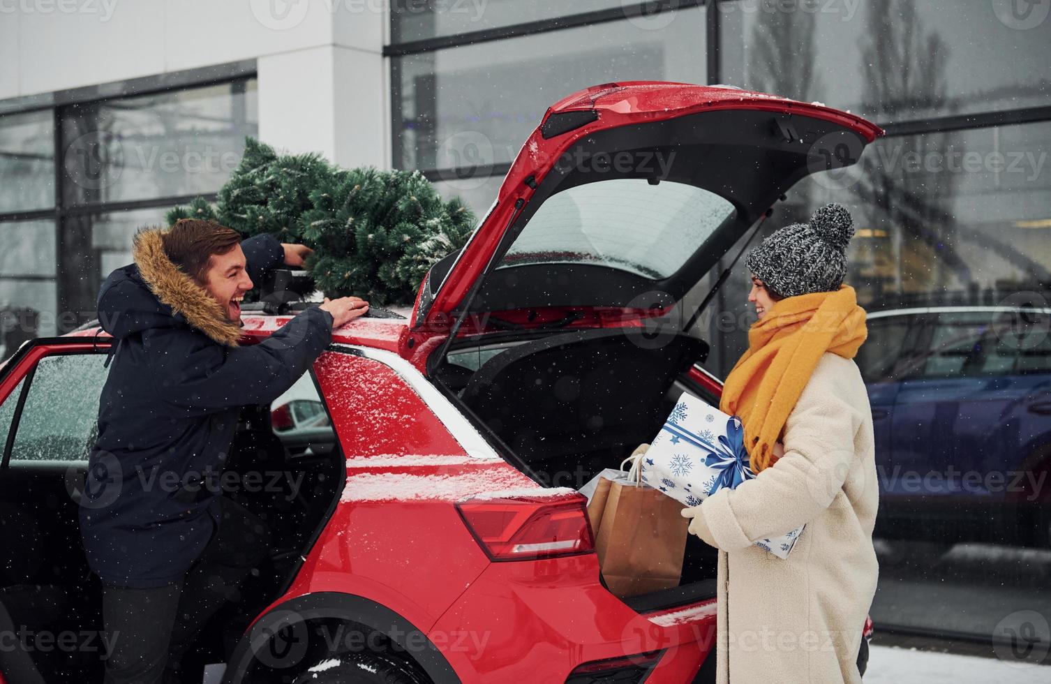 giovane coppia con regalo scatole è vicino auto con albero su il superiore. insieme all'aperto a inverno tempo foto