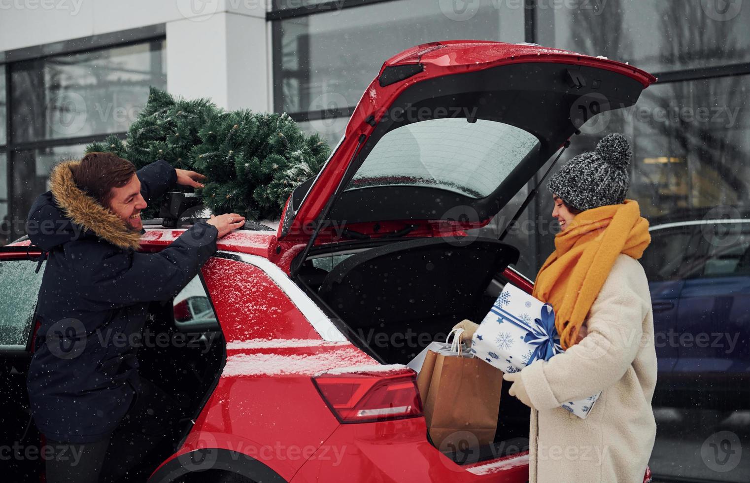 giovane coppia con regalo scatole è vicino auto con albero su il superiore. insieme all'aperto a inverno tempo foto