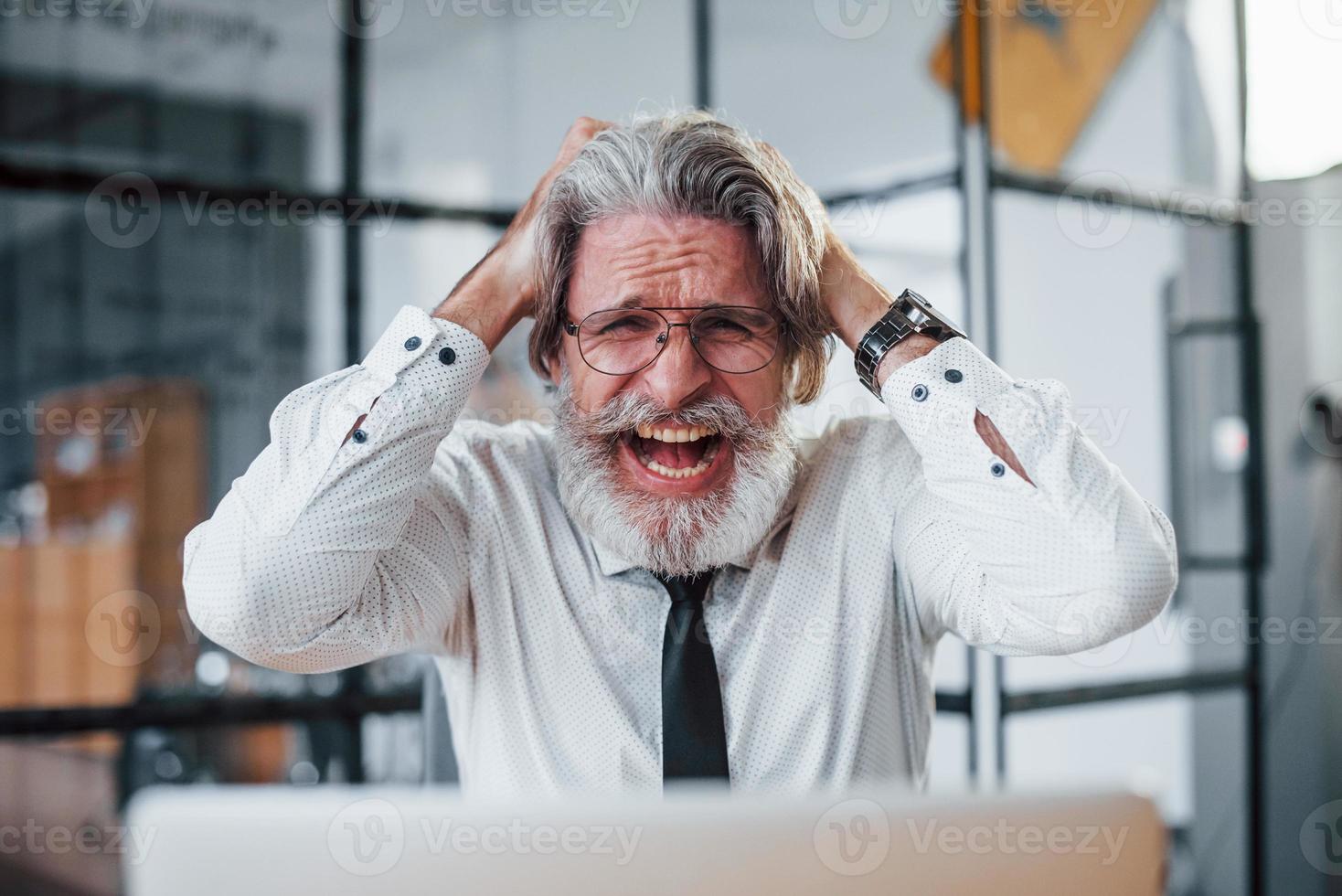 espressive maturo uomo d'affari con grigio capelli e barba nel formale Abiti è nel il ufficio contro il computer portatile foto