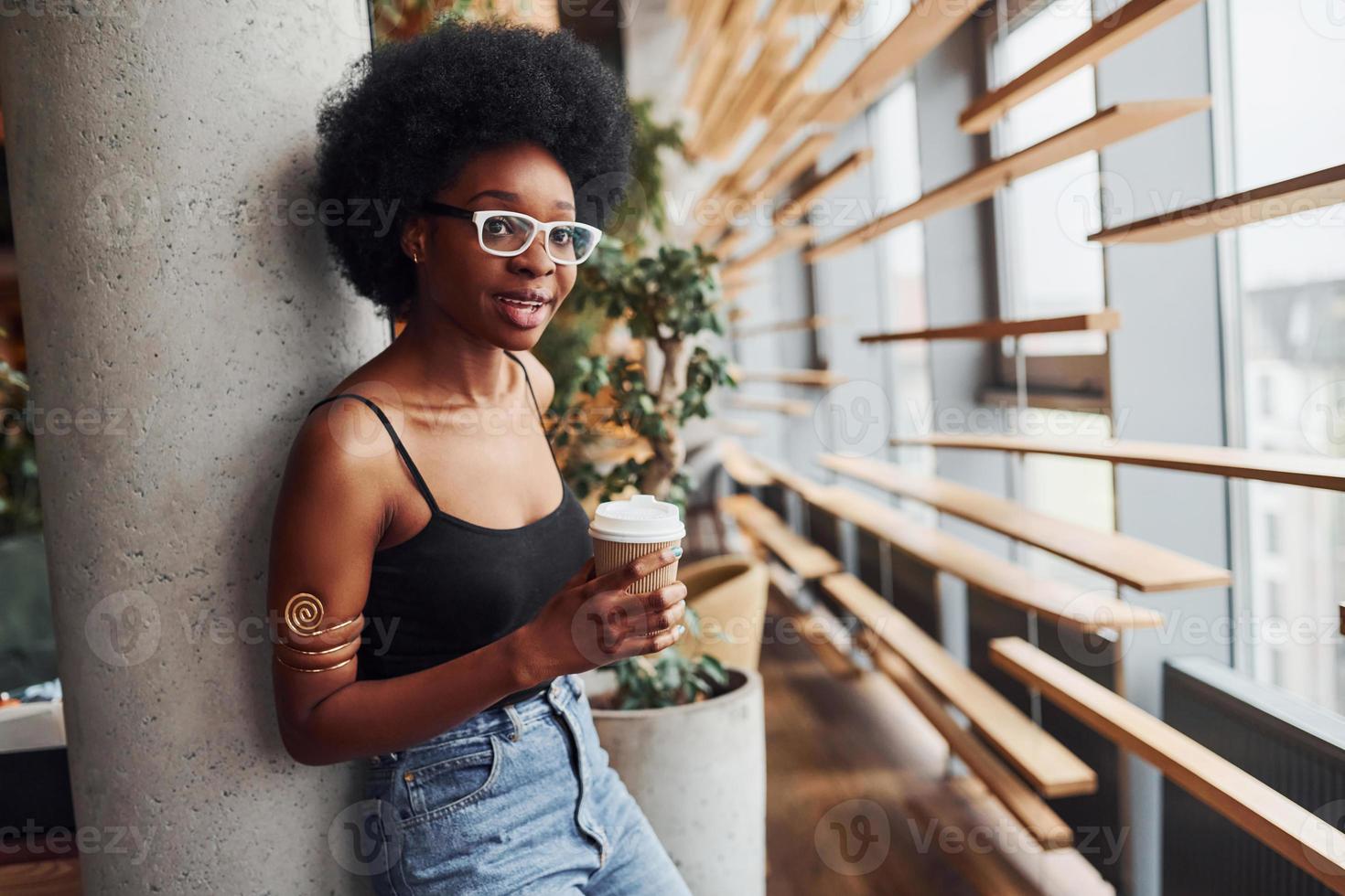 africano ragazza con Riccio nero capelli e nel casuale Abiti in piedi in casa con tazza di bevanda foto