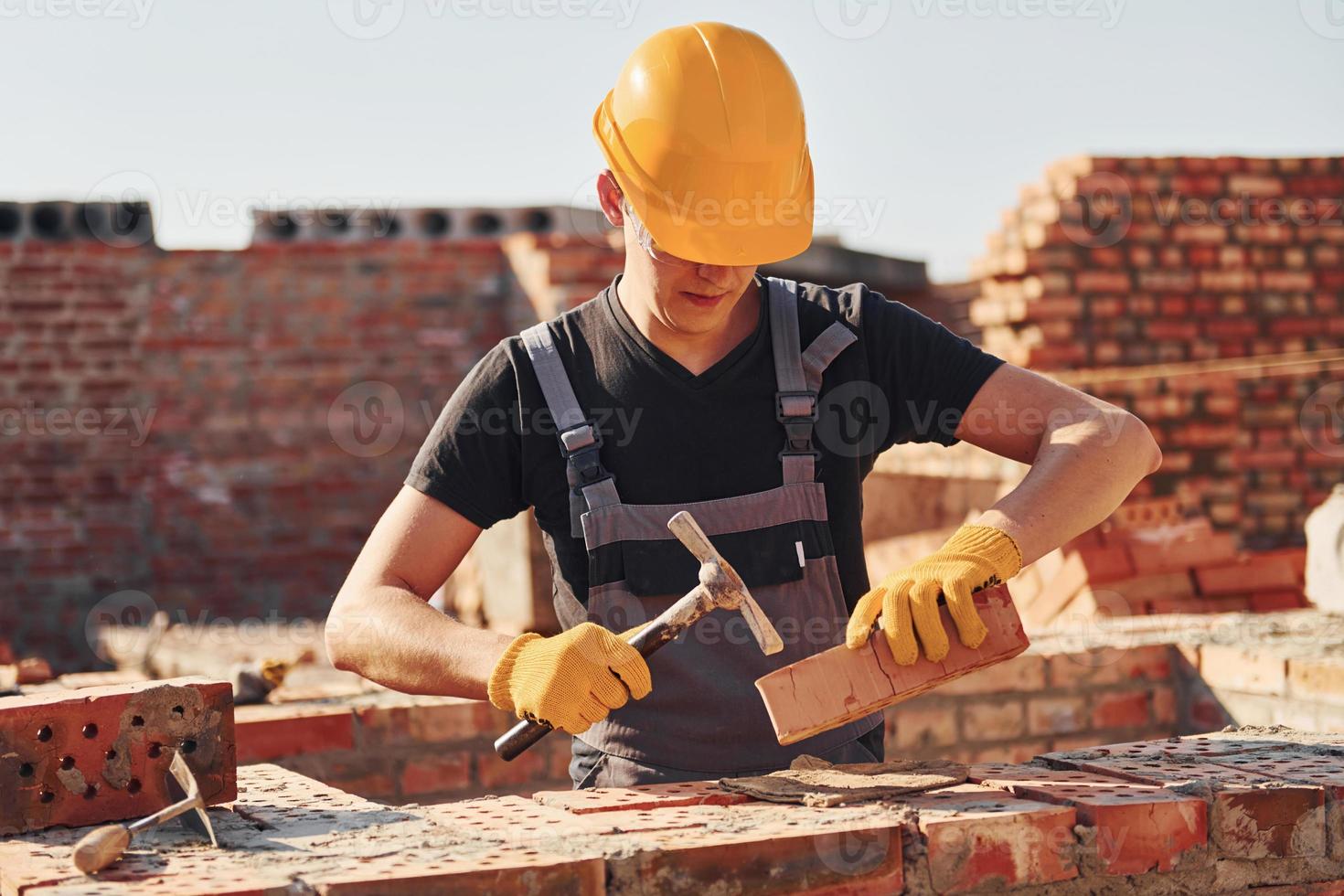 Tenere mattone e utilizzando martello. costruzione lavoratore nel uniforme e sicurezza attrezzatura avere lavoro su edificio foto