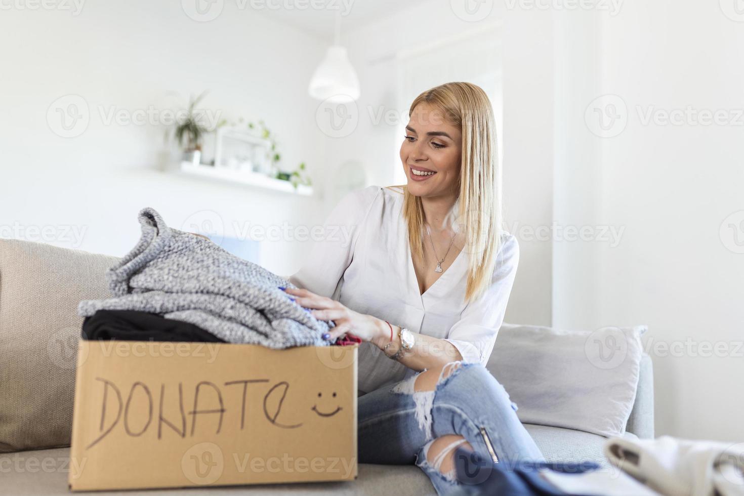 giovane donna seduta su divano preparazione pacco per invio per bisognoso umano. ragazza con grande genere cuore mette Usato Abiti nuovo indossare nel donazione scatola, concetto di cura di senza casa persone foto