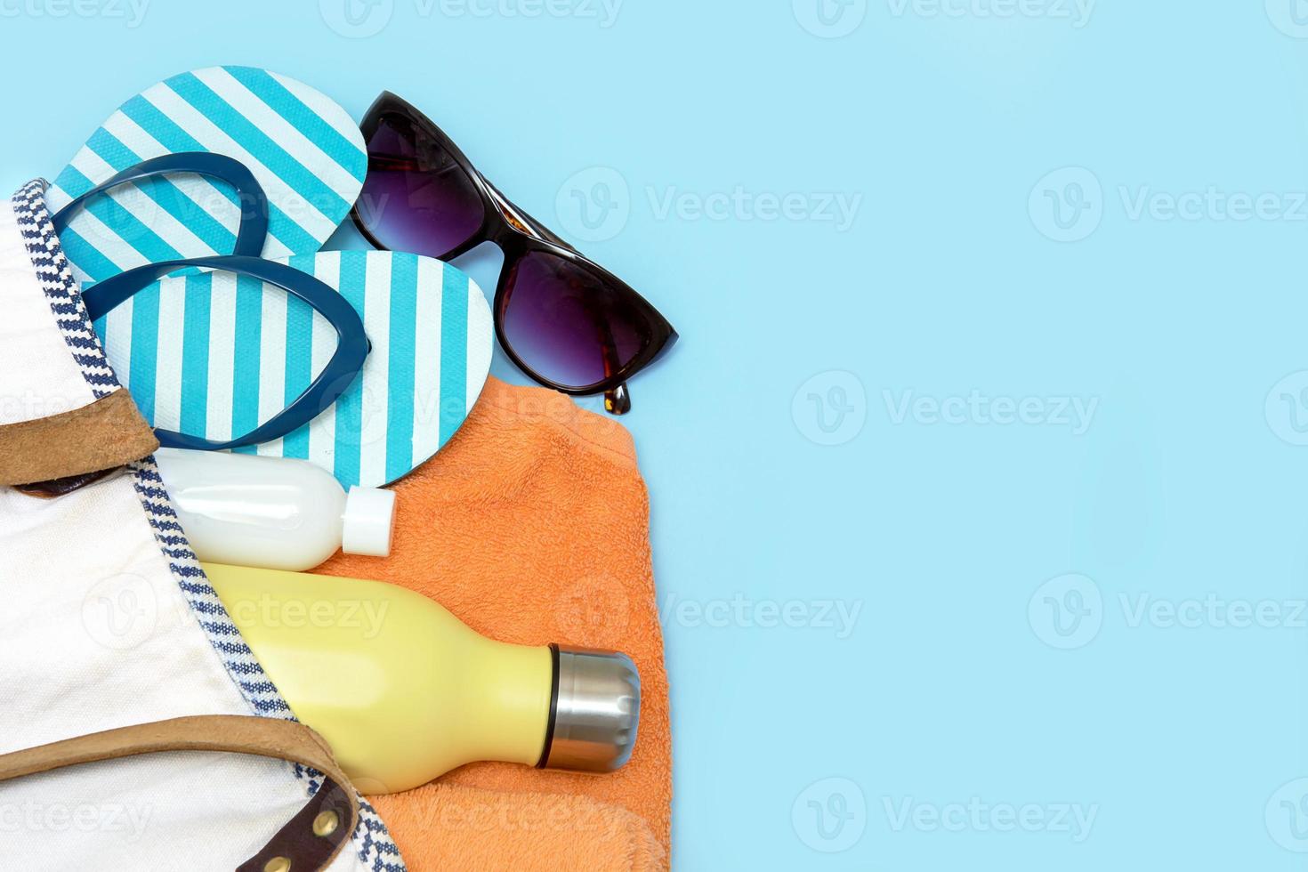 spiaggia Accessori - un' bottiglia con un' morbido bere, sole crema, occhiali da sole, Flip flop, un' asciugamano nel un' spiaggia Borsa su un' blu sfondo con copia spazio foto