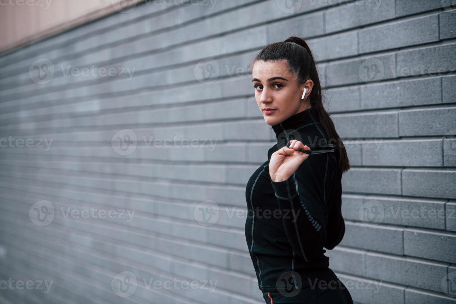 giovane allegro ragazza nel nero abbigliamento sportivo in piedi con saltare corda nel mani all'aperto vicino grigio parete foto