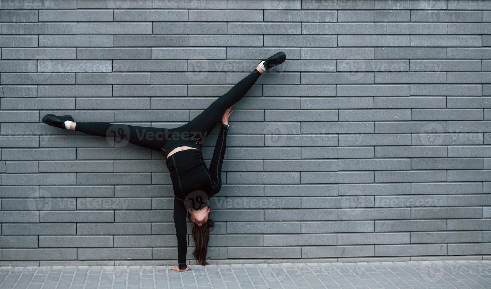 giovane allegro ragazza nel nero abbigliamento sportivo fare difficile handstand esercizi all'aperto vicino grigio parete foto