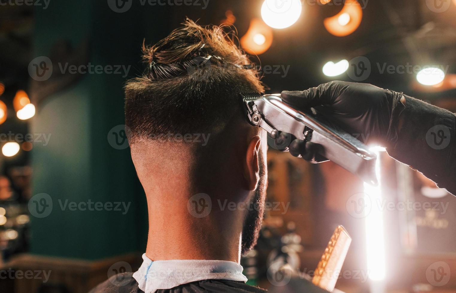 vicino su Visualizza di giovane barbuto uomo quello seduta e ottenere taglio di capelli nel barbiere negozio foto