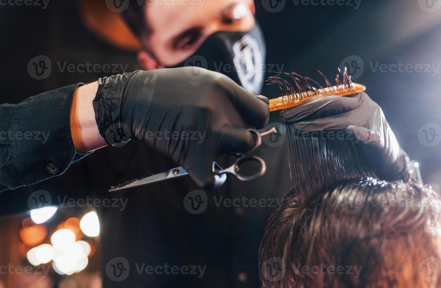 giovane barbuto uomo seduta e ottenere taglio di capelli nel barbiere negozio di tipo nel nero protettivo maschera foto