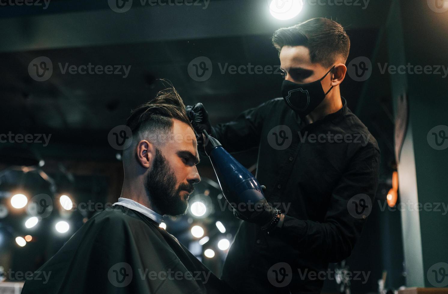 giovane barbuto uomo seduta e ottenere taglio di capelli nel barbiere negozio di tipo nel nero protettivo maschera foto