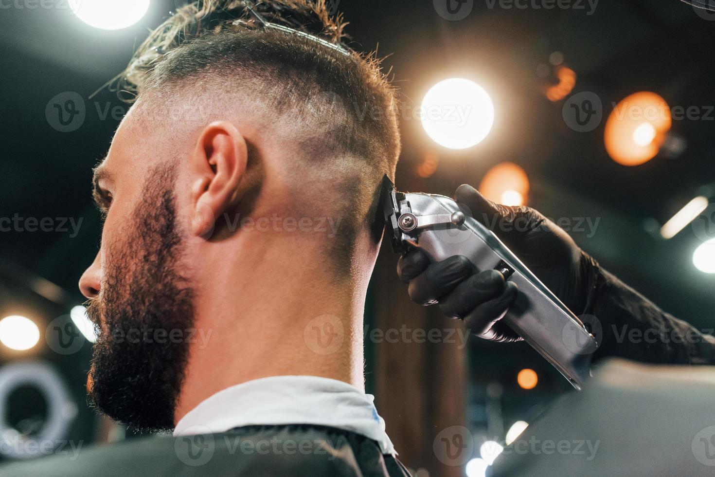 vicino su Visualizza di giovane barbuto uomo quello seduta e ottenere taglio di capelli nel barbiere negozio foto