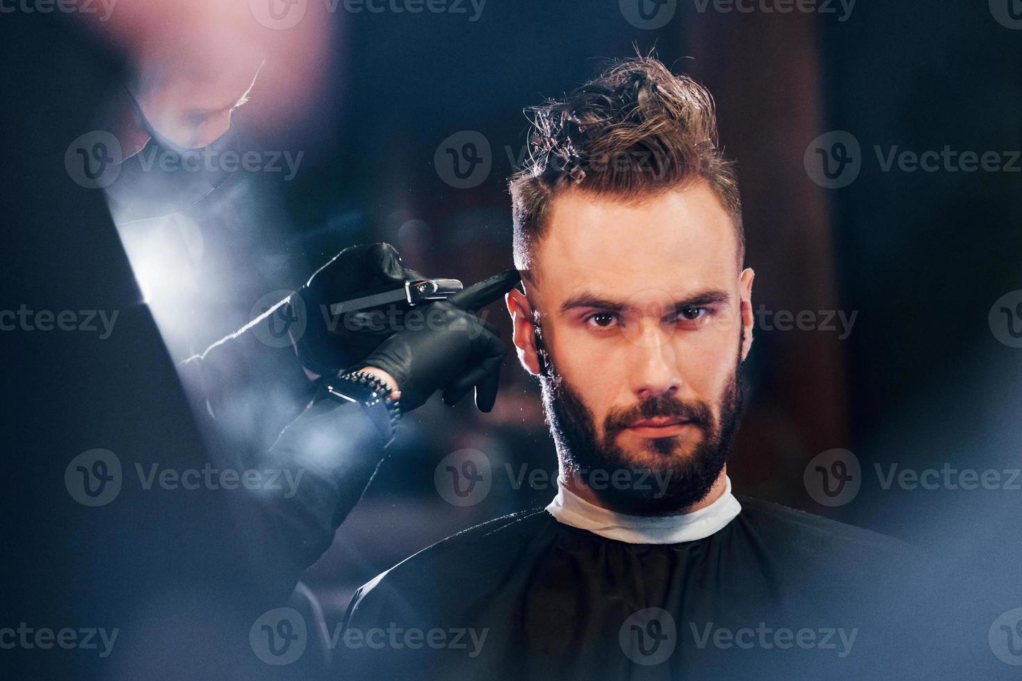giovane barbuto uomo seduta e ottenere taglio di capelli nel barbiere negozio foto