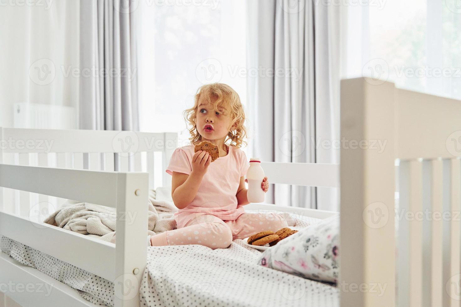 seduta su il letto, mangiare biscotti e potabile latte. carino poco ragazza nel casuale Abiti è in casa a casa a giorno foto