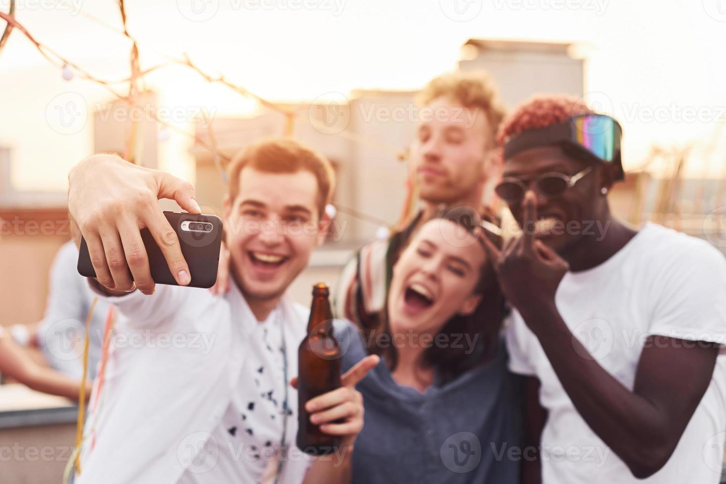 in piedi insieme e assunzione autoscatto. gruppo di giovane persone nel casuale Abiti avere un' festa a tetto insieme a giorno foto