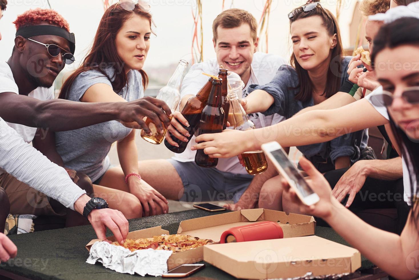 delizioso Pizza. gruppo di giovane persone nel casuale Abiti avere un' festa a tetto insieme a giorno foto