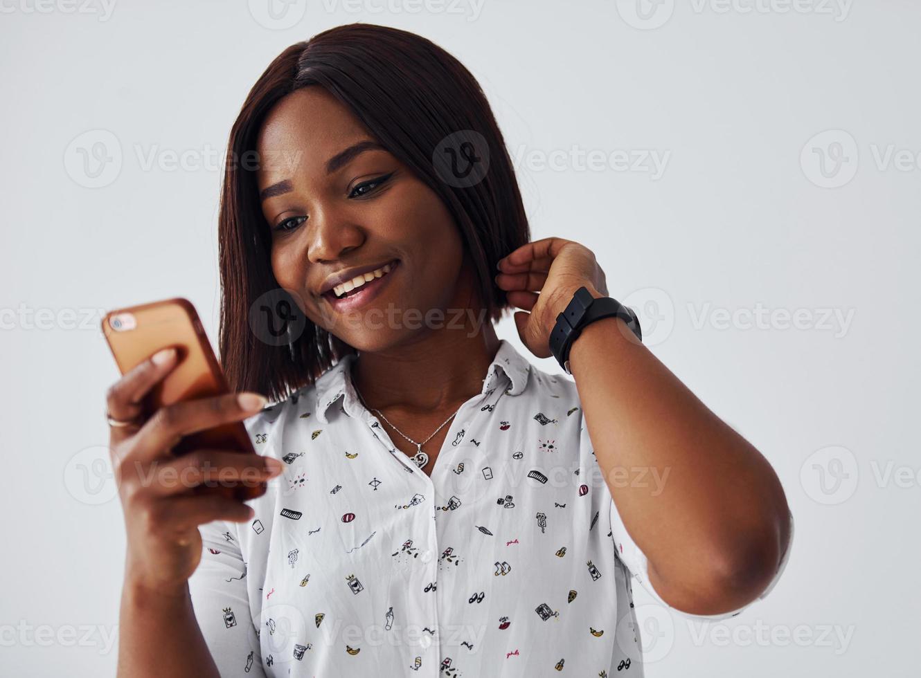 positivo africano americano donna nel bianca camicia in piedi in casa contro bianca parete con Telefono nel mano foto