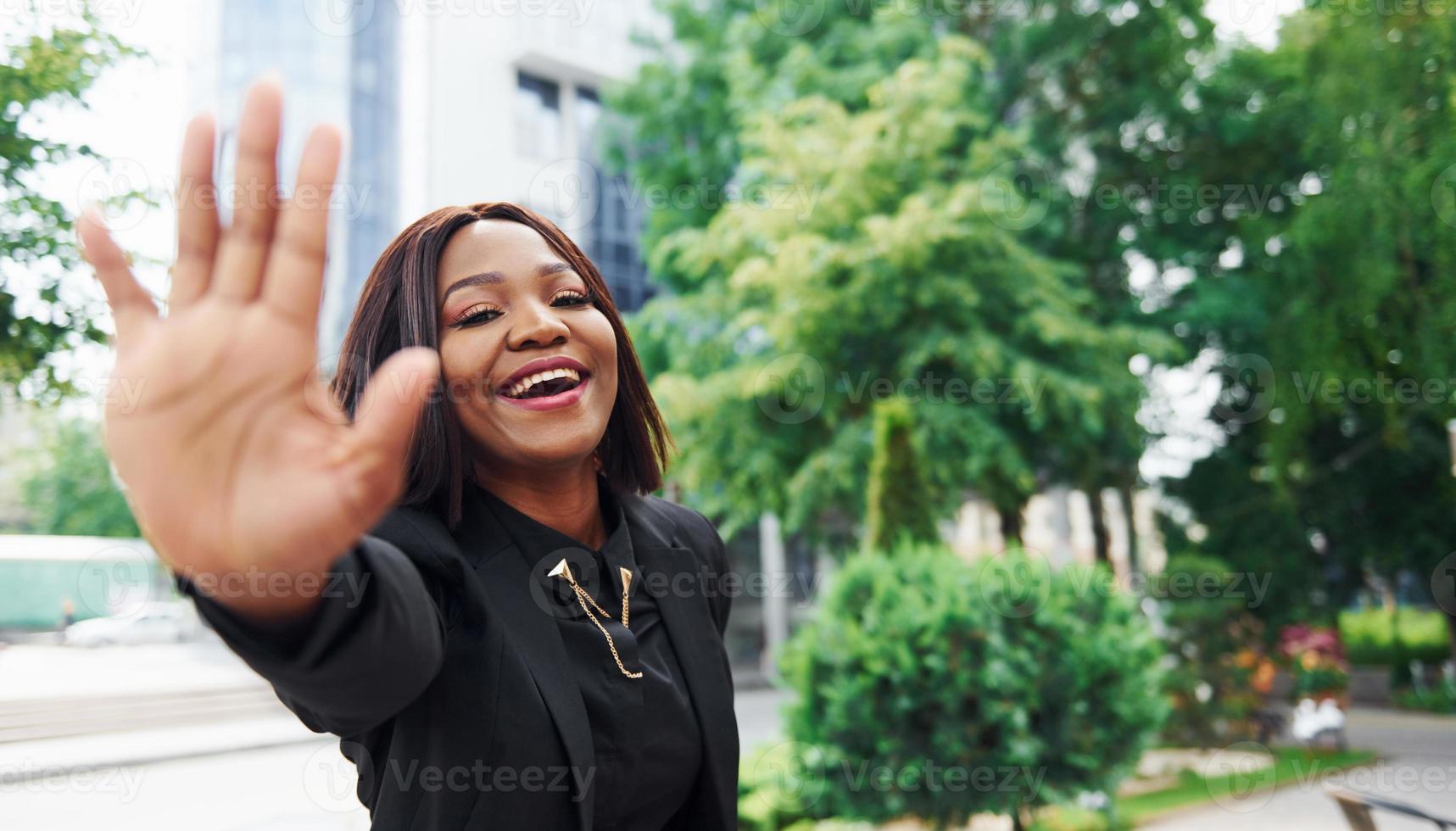 alto cinque gesto. giovane afro americano donna nel alla moda Abiti all'aperto nel il città vicino verde alberi e contro attività commerciale edificio foto