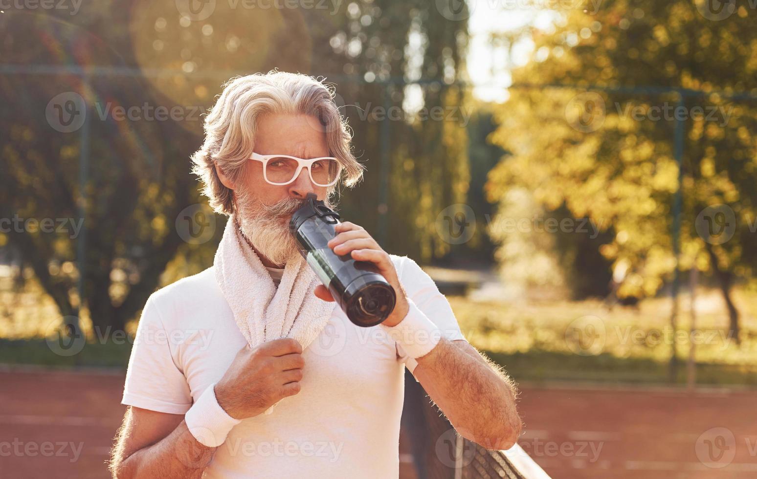 assunzione un' rompere e potabile acqua. anziano elegante uomo nel bianca camicia e nero allegro pantaloncini su tennis Tribunale foto