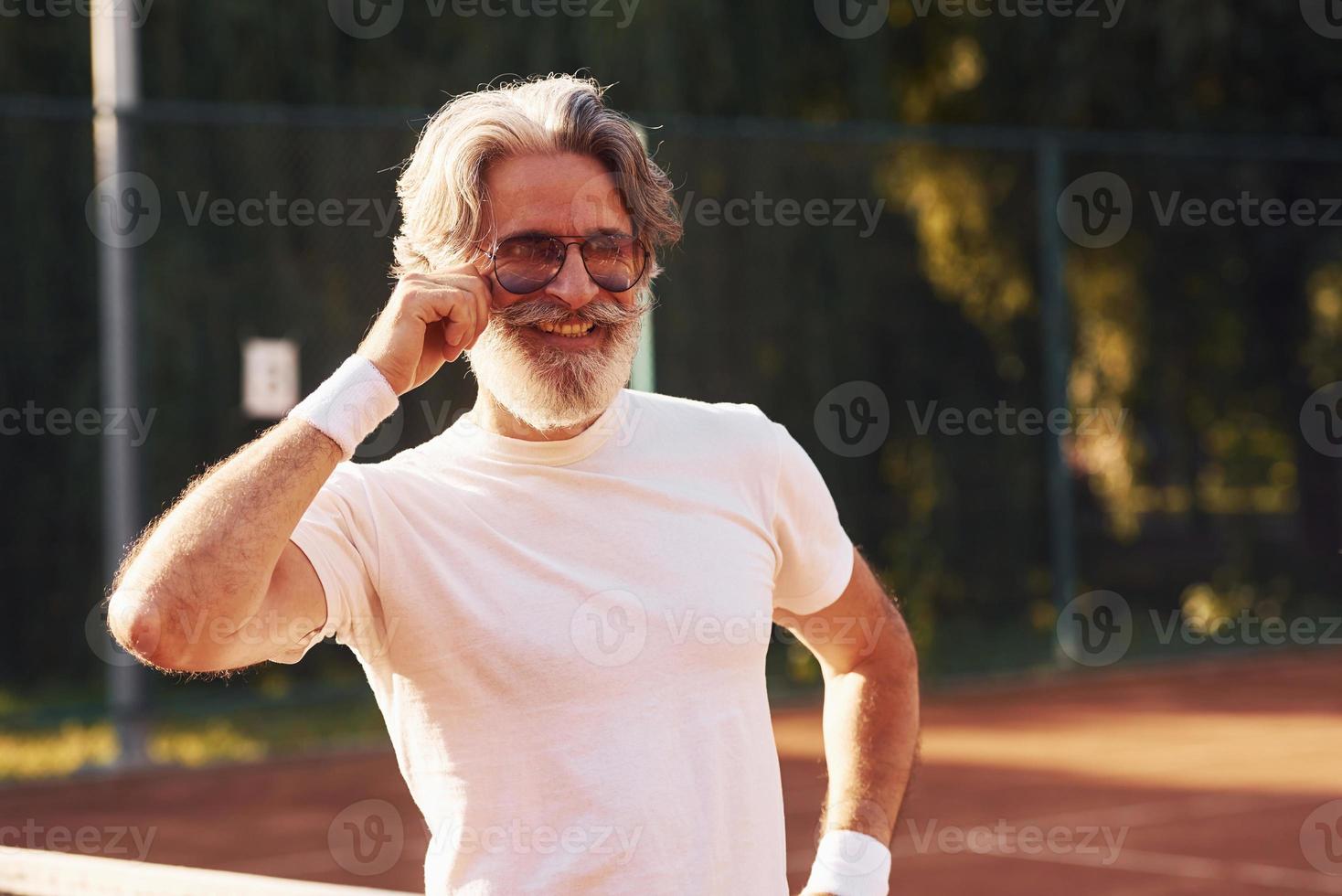 anziano elegante uomo nel occhiali, bianca camicia e nero allegro pantaloncini su tennis Tribunale foto