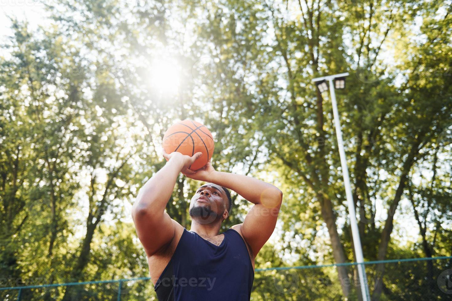 bellissimo verde alberi su sfondo. africano americano uomo giochi pallacanestro su il Tribunale all'aperto foto