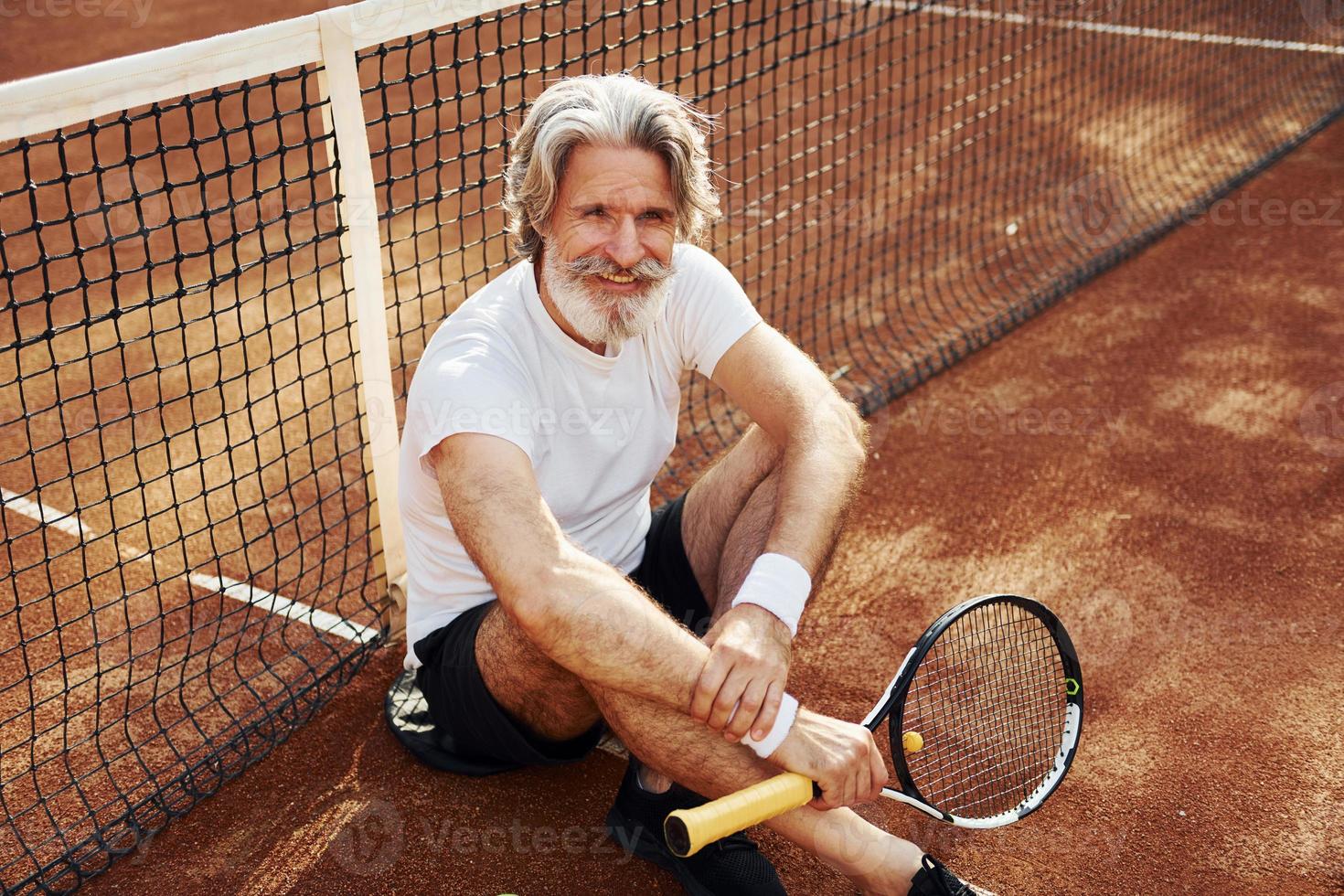 si siede su il terra e prende rompere. anziano moderno elegante uomo con racchetta all'aperto su tennis Tribunale a giorno foto