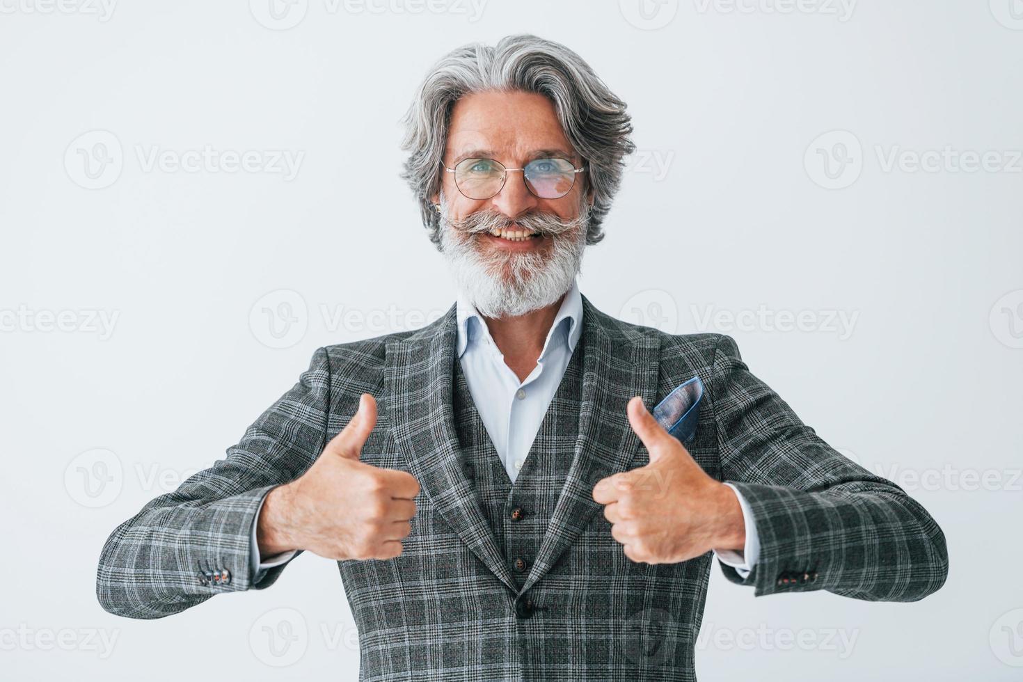 nel elegante Abiti. in piedi contro bianca parete. anziano elegante moderno uomo con grigio capelli e barba in casa foto